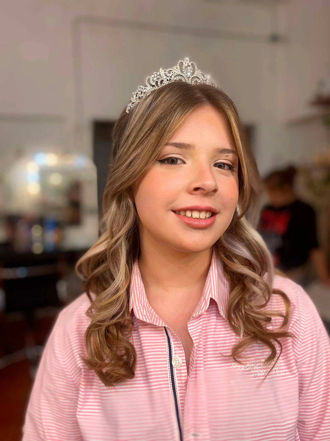 Peinado infantil de ondas sueltas y rizos definidos en cabello castaño claro, con un acabado brillante y profesional para el sitio de Meli Figarola. El look destaca la textura y el volumen del cabello, realzado por una tiara de cristales y una sonrisa alegre en un entorno de preparación cálidamente iluminado.
