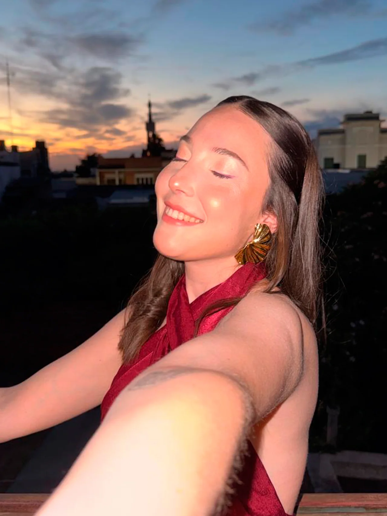 Peinado de cabello largo y liso en tono castaño con acabado sedoso y saludable para el sitio de Meli Figarola. El look social destaca el brillo natural del cabello bajo la luz del atardecer, peinado sutilmente detrás de la oreja para resaltar los accesorios y un maquillaje luminoso.