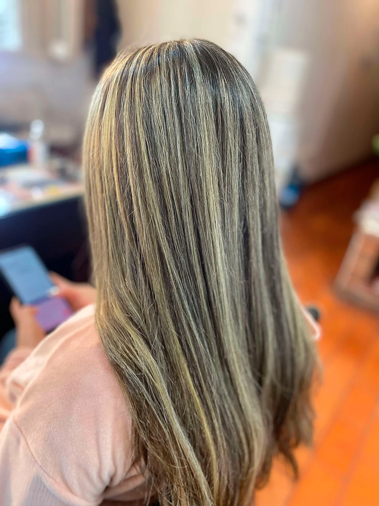 Primer plano de la vista trasera del cabello largo y liso de una mujer, que presenta mechas rubias finas y reflejos multi-tonales sobre una base castaña oscura. Viste una prenda de color rosa claro. El fondo desenfocado muestra un interior con suelo de madera y una persona con un smartphone