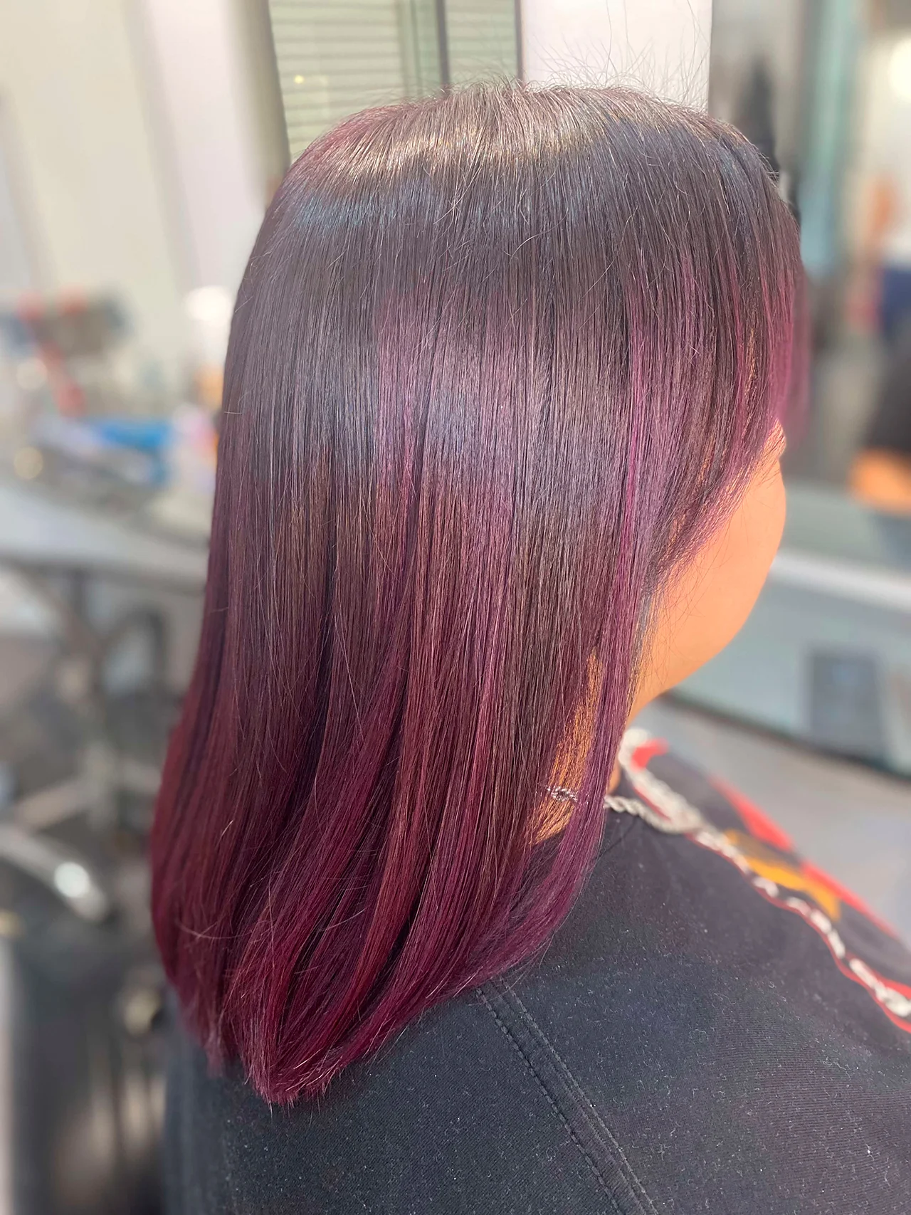 Un primer plano lateral de un corte bob de longitud media y liso de una persona, mostrando una coloración fantasía vibrante. El cabello tiene una base oscura con reflejos intensos y multidimensionales en tonos ciruela, magenta y violeta, con puntas de color ciruela más saturadas. El cabello está brillante y bien peinado. La persona lleva una camiseta negra y una cadena plateada. El fondo es un salón de belleza desenfocado con sillas de salón y espejos.