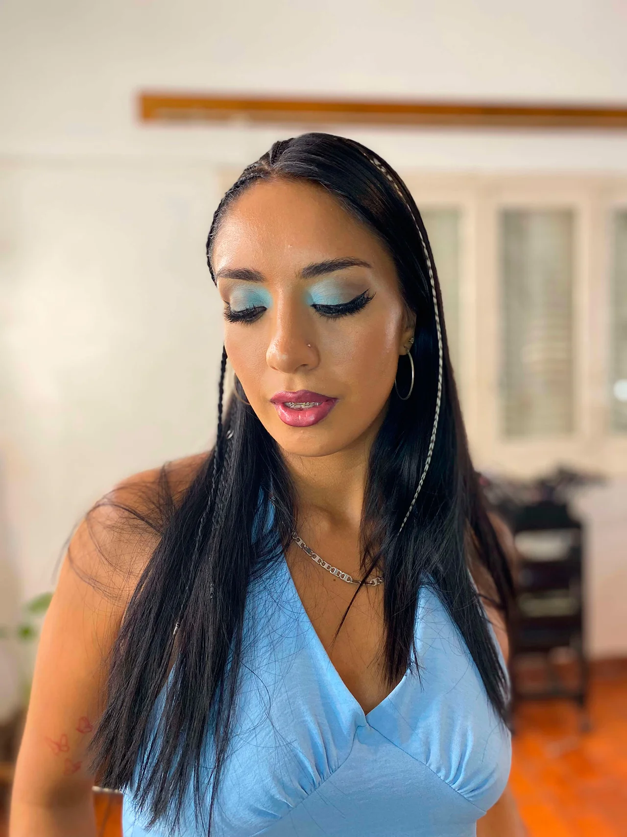 Un primer plano de una mujer joven con los ojos cerrados, mostrando un maquillaje de ojos azul claro vibrante con delineado alado negro y pestañas postizas. Su cabello negro largo tiene trenzas laterales con cuentas. Ella tiene labios fucsia brillantes, usa pendientes de aro y un collar de cadena delicado, y viste un top halter de color azul claro en un interior desenfocado.