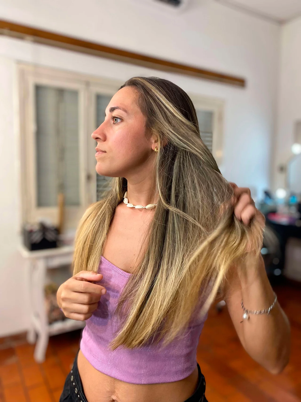 Retrato de una mujer joven con el cabello largo y liso con mechas rubias. Ella está de pie en un interior, pasando sus manos por su cabello con una expresión serena mientras mira hacia un lado. Lleva un collar de caracoles, un top corto de punto acanalado en color lavanda y pantalones oscuros. El fondo muestra un ambiente hogareño con suelo de madera y una ventana con marcos blancos bajo una luz natural suave.