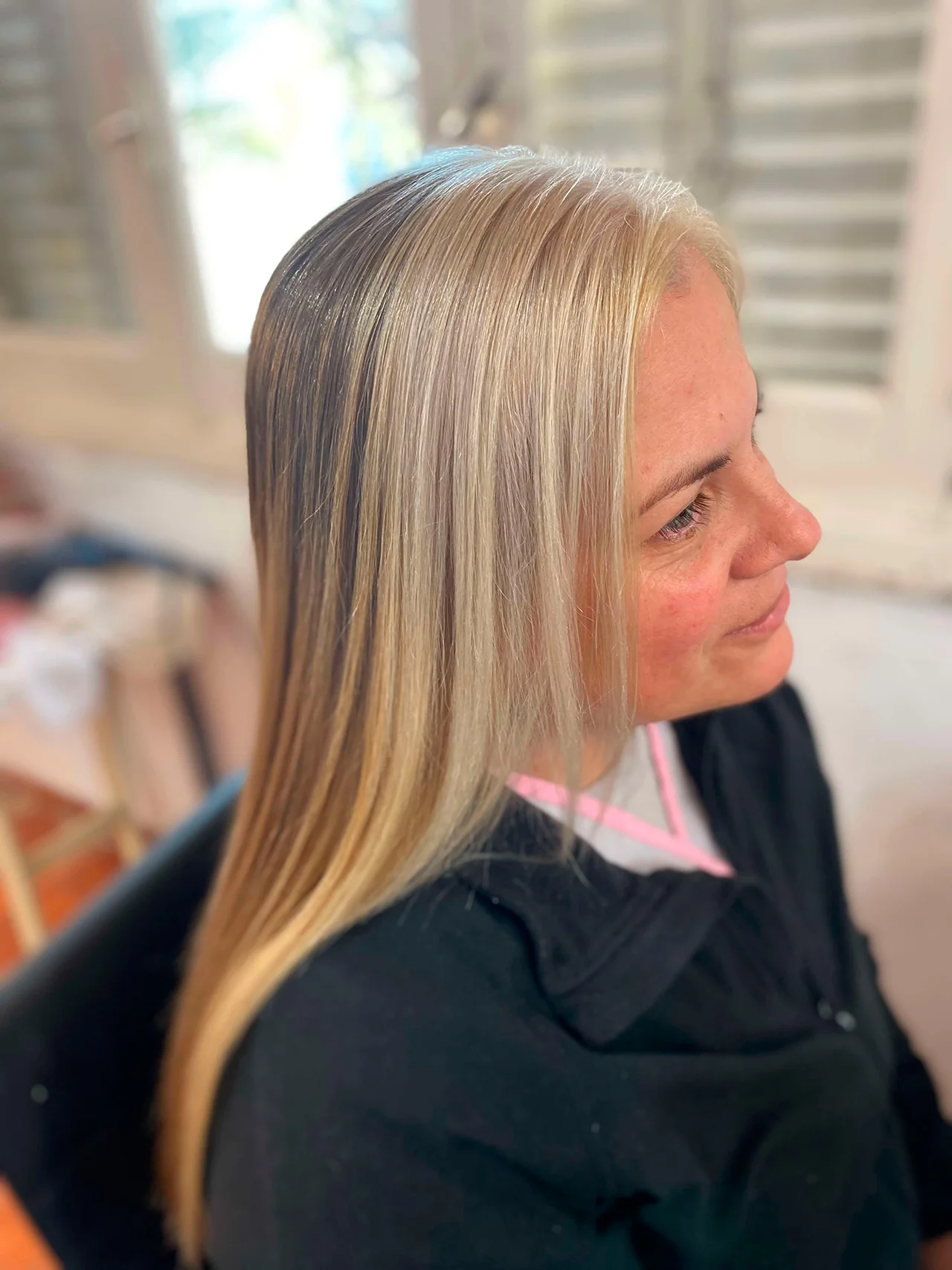 Perfil lateral de una mujer con cabello largo y liso en tonos rubios. Destaca una sección frontal en rubio platino que se funde con reflejos más cálidos y miel hacia la parte posterior. La mujer sonríe levemente y viste una chaqueta negra sobre una prenda blanca con detalles rosados. El fondo muestra un interior luminoso con persianas de madera.