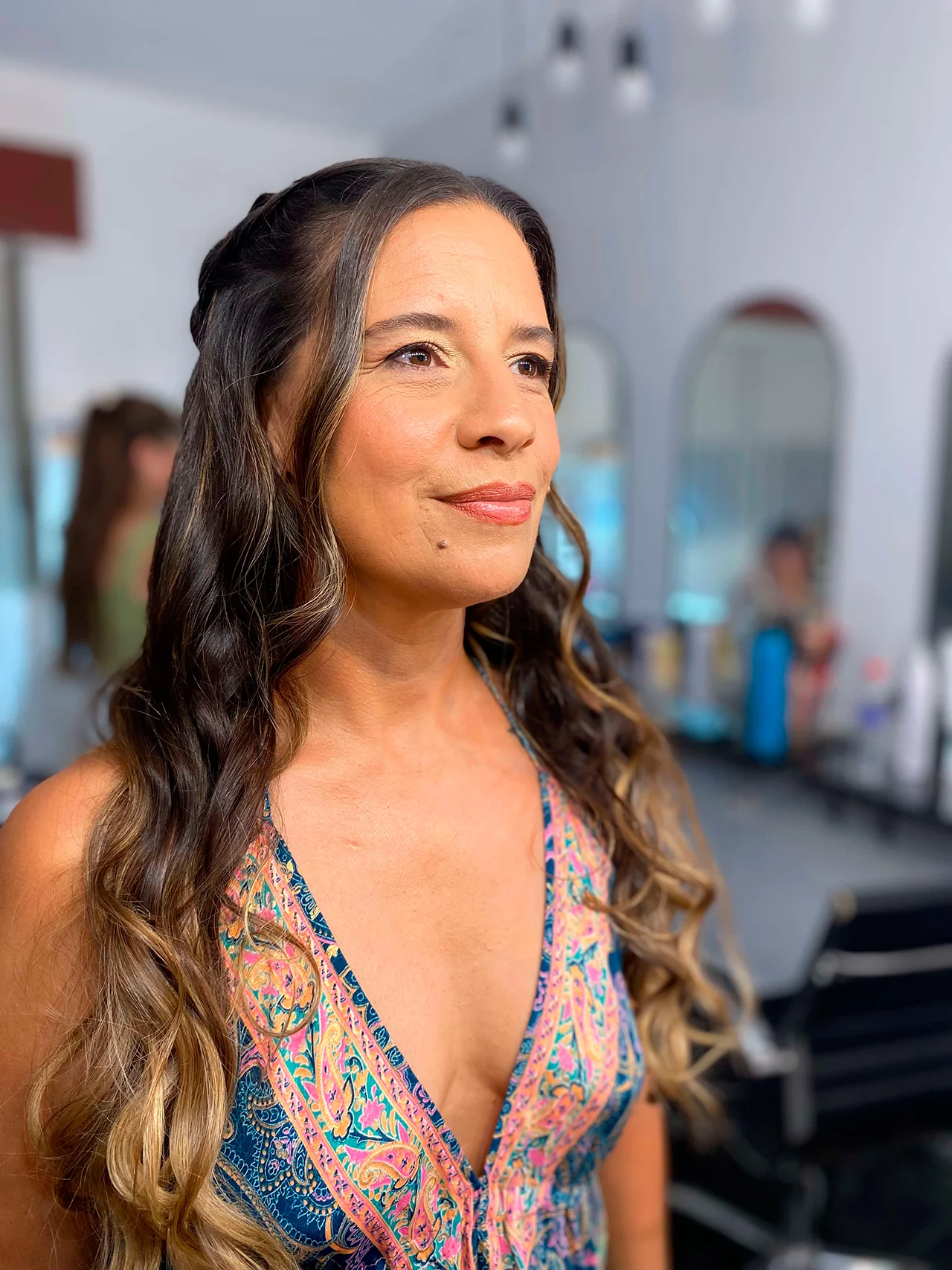 Retrato de una mujer con el pelo largo oscuro y puntas rubias balayage, peinado con un semi-recogido con una trenza en la parte superior y ondas sueltas y fluidas. Lleva un maquillaje de aspecto natural y un vestido de tirantes con un patrón de cachemira vibrante y colorido. El fondo es el interior desenfocado de un salón de belleza con espejos de arco y productos.
