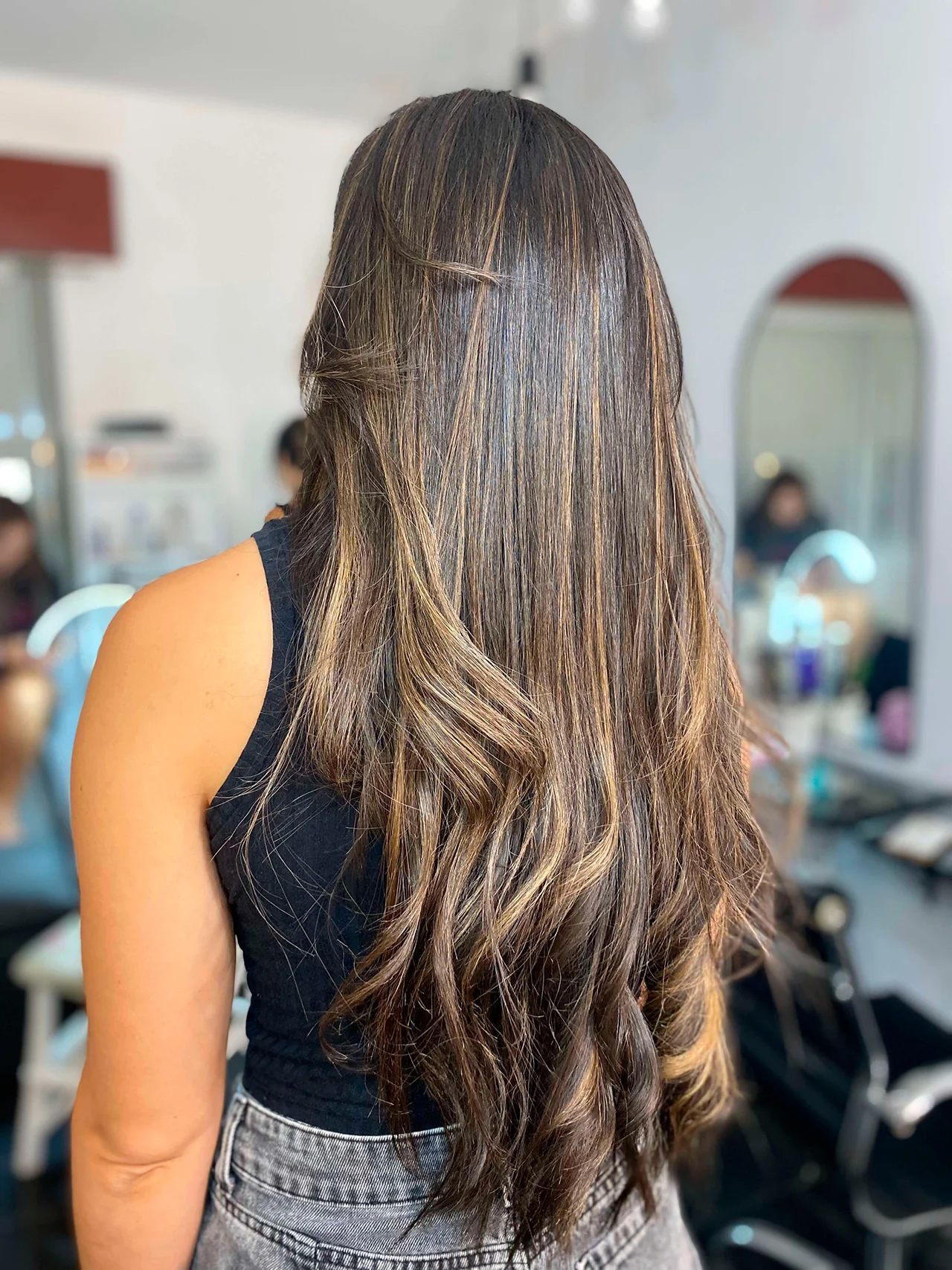Vista trasera de una mujer con el cabello largo y ondulado, que presenta una técnica de balayage dimensional con reflejos en tonos miel y rubio claro sobre una base castaña media. Las raíces están suaves y el final tiene ondas sueltas y fluidas. Lleva una camiseta negra de tirantes y está en un salón de belleza con un fondo borroso de espejos y equipos de peluquería.